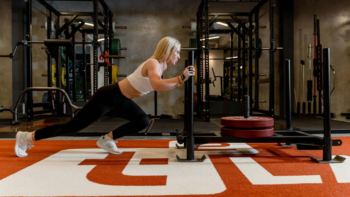 Athlete driving a weighted sled across gym turf with the back leg fully extended — the heavy calf push-off that can strain a calf in HYROX training in Tampa.