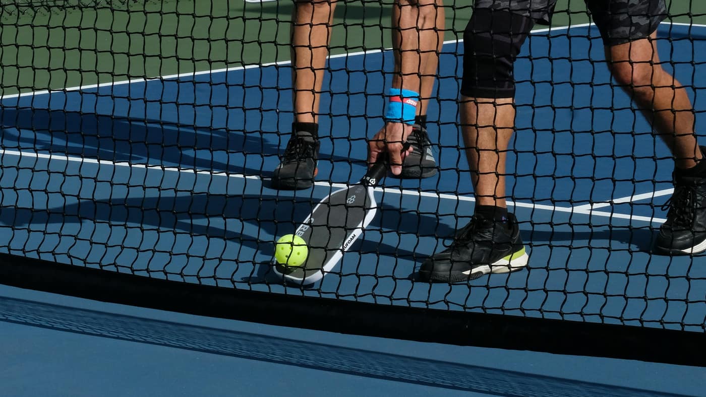 Two pickleball players at the net, calves and court shoes in view as one reaches low with the paddle — the push-off and lunge behind “tennis leg” calf strains in Tampa Bay court athletes.