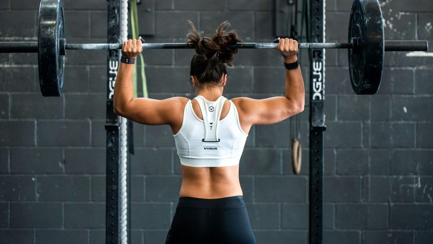 Lifter pressing a barbell overhead in a rack, seen from behind with both shoulder blades working — the overhead pressing where shoulder pain in Tampa lifters often shows up first.