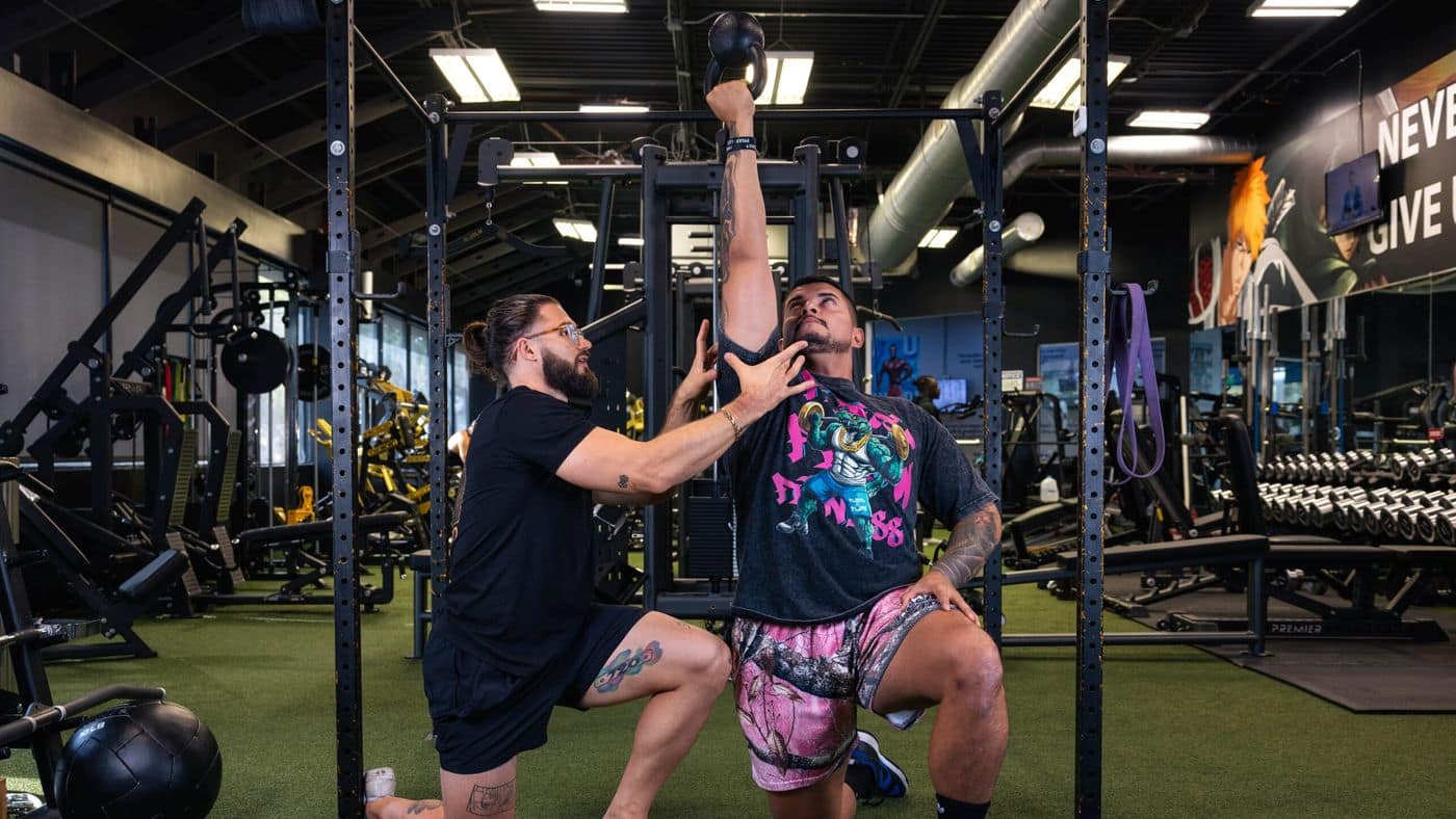 Forward Physio doctor of physical therapy coaching a lifter through a half-kneeling bottoms-up kettlebell press in a Tampa gym — overhead control work used in shoulder rehab for lifters.
